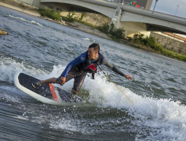 A man in a blue and black wet suit rides on a wake board. A bridge can be seen in the background