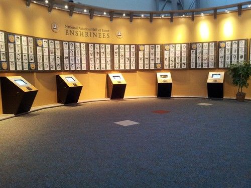 A photo of a wall with the words National Aviation Hall of Fame Enshrinees. On the wall are the photos of enshrinees and in front of the wall are  four information kiosks.