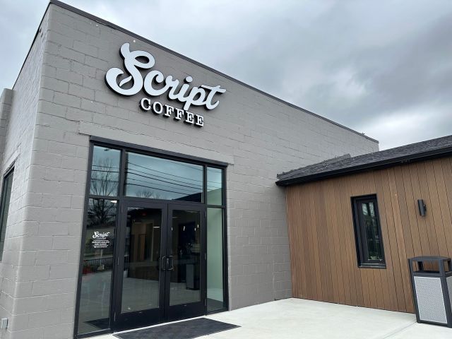 Photo of Script Coffee store front. A tall, brick building that has been painted grey with a set of doors. Above the doors are the words Script Coffee. To the right, a smaller addition has been attached to the building. It is brown with a black roof. A public trash can sits against the wall.