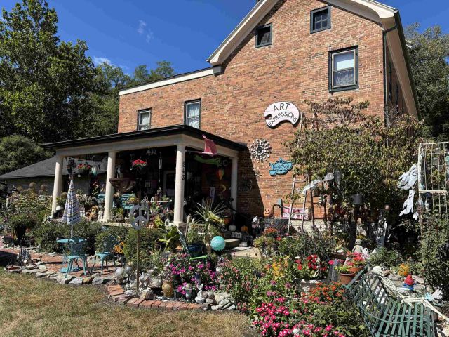 Photo of Art Expressions store front. A large brick building with a sign shaped like a large paint palette hangs from the wall. It says Art Expressions. A ladder is propped against the wall nearby. The surrounding yard is full of art pieces, small statues, and wind chimes. A table and two chairs are set up in the background, and a public bench is in the foreground.