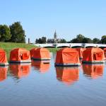 Eleven orange floating tents sit on top of the water on a clear, sunny day. A white bridge and a domed white building can be seen in the background.